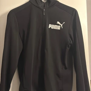 Svart träningsjacka från Puma - Snygg svart träningsjacka från Puma med dragkedja och logga på bröstet. Perfekt för träning eller som en sportig vardagslook. Jackan har långa ärmar och en hög krage för extra komfort.