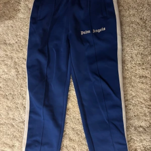 Blå Palm Angels byxor - Snygga blå Palm Angels byxor med vita sidopaneler och logotyp på framsidan. Byxorna har en elastisk midja för extra komfort och en stilren design som passar perfekt för en avslappnad look.