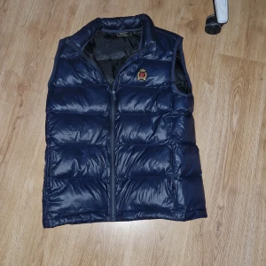 Mörkblå dunväst - Säljer en snygg mörkblå dunväst från SikSilk. Västen har en stilren design med ett litet emblem på bröstet. Perfekt för kyligare dagar när du vill hålla stilen. Riktigt go väst