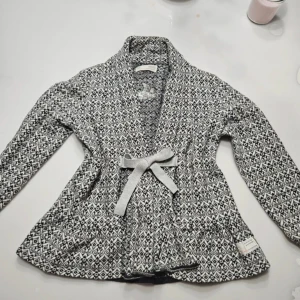 Odd Molly kofta - Snygg svartvit mönstrad kofta från Odd Molly med rosett i midjan. Koftan har långa ärmar och ett elegant mönster som ger en stilren look. Perfekt för att hålla sig varm och chic.