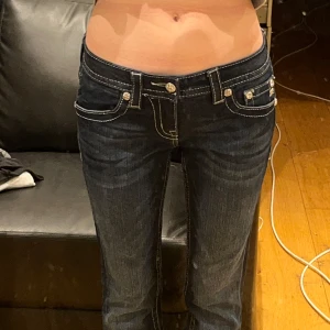 Mörkblå jeans med detaljer från Miss Me - Snygga mörkblå jeans från Miss Me med unika detaljer. Byxorna har låg midja och är dekorerade med glittriga paljetter och broderier på bakfickorna. Perfekta för att ge din outfit en extra touch. Bootcut. Midja: 34, Innerben 82. 