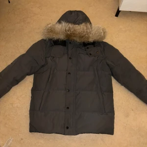 Grå dunjacka från Canada Goose - Säljer en grå dunjacka från Canada Goose med pälskantad huva. Jackan har flera praktiska fickor och stängs med knappar. Perfekt för kalla vinterdagar. Den är inte så mörk grå som på första bilden det va dåligt ljus färgen syns som bäst på andra bilen. Skriv vid frågor. 