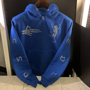 Blå rihnestone zip hoodie - Snygg blå hoodie med dragkedja och glittriga detaljer i form av skelett och symboler. Perfekt för att sticka ut med en unik stil. Hoodien har långa ärmar och en bekväm passform samt är passformen på luvan perfekt. Hör av er vid minsta fundering🙌