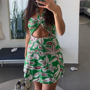 Grön blommig klänning från Zara - Trendig grön klänning med blommigt mönster och knytning framtill. Den har en öppen rygg och är ärmlös, vilket gör den perfekt för varma dagar.