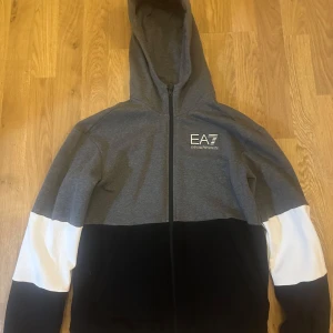 Grå och svart huvtröja från EA7 - Snygg huvtröja från EA7 i grått och svart med vita detaljer. Tröjan har en dragkedja framtill och en luva. Perfekt för en avslappnad stil. Passar storlek s