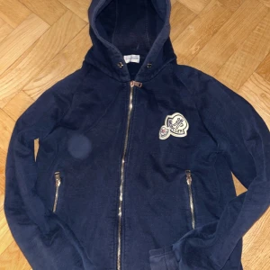 Mörkblå hoodie från Moncler - Snygg mörkblå hoodie från Moncler med dragkedja och huva. Den har ett broderat Moncler-märke på bröstet och praktiska fickor med dragkedjor. Perfekt för en avslappnad stil. Inga defekter eller skador alls! Är storlek S men passar M också