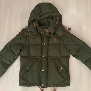 Grön dunjacka från Polo Ralph Lauren - Säljer en snygg grön dunjacka från Polo Ralph Lauren med huva och dragkedja. Jackan har knappar och praktiska fickor framtill. Perfekt för kyliga dagar!💕❄️💋pris kan absolut diskuteras 