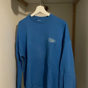 Blå sweatshirt från Junkjard  - Säljer en blå sweatshirt från Junk jard med rund hals och ett litet tryck på bröstet. Tröjan har långa ärmar och en avslappnad passform. Perfekt för en casual look!