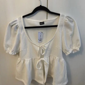 Vit blus med puffärmar från Gina Tricot - Charmig vit blus från Gina Tricot med puffiga kortärmar och knytning framtill. Blusen har en luftig passform och är perfekt för varmare dagar. Den har en söt volangdetalj vid midjan som ger en romantisk touch.