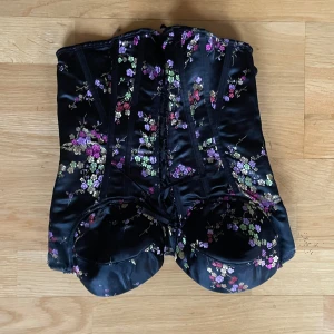 Svart blommig korsett - Elegant svart korsett med blommigt mönster i lila, rosa och grönt. Tillverkad i ett glansigt material med snörning bak för justerbar passform. Perfekt för att ge en stilfull touch till din outfit. Passar om du har stora bröst!! 