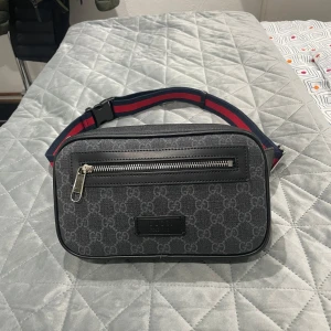 Svart midjeväska från gucci - Snygg svart midjeväska från gucci med diskret mönster och justerbart band i rött och blått. Väskan har en praktisk dragkedja framtill och är perfekt för att hålla dina saker säkra och nära till hands.