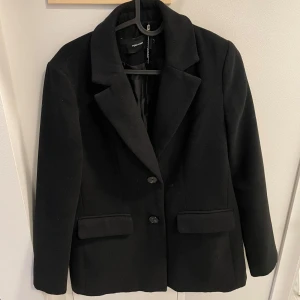 Svart kavaj från Vero Moda - Elegant svart kavaj från Vero Moda med klassisk design. Kavajen har långa ärmar och knäpps med två knappar framtill. Perfekt för en stilren look. Tyget är tjockare som en kappa 