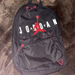 Svart ryggsäck från Air Jordan - Snygg svart ryggsäck från Jordan med det ikoniska röda logotyptrycket. Ryggsäcken har en praktisk framficka med dragkedja och justerbara axelremmar för bekväm passform. Perfekt för vardagsbruk, träning eller skola. Använd men finns inga märken på slitage. Köpt för 600kr på zalando