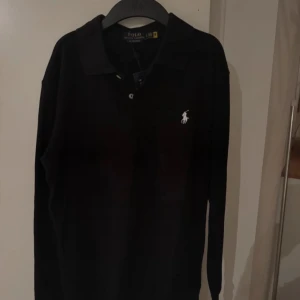 Svart pikétröja från Ralph Lauren lappen sitter kvar  - Säljer en stilren svart pikétröja från Ralph Lauren med lång ärm och klassisk krage. Tröjan har en broderad logotyp på bröstet och är i en custom slim fit passform. Perfekt för en elegant och avslappnad look.