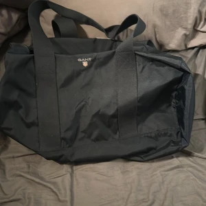 Svart väska från Gant - Säljer en stilren svart väska från Gant. Perfekt för träning eller som weekendbag. Väskan har en praktisk dragkedja och robusta handtag för enkel bärning. Den är rymlig och tillverkad i slitstarkt material. Den har aldrig varit använd då jag inte gillat den å tyckt att den inte passar min still