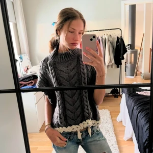 Grå stickad väst med flätmönster - Säljer en mysig grå stickad väst med flätmönster och dekorativa tofsar i nederkant. Perfekt för kyliga dagar och ger en avslappnad look. Passar bra över en enkel topp eller tröja.