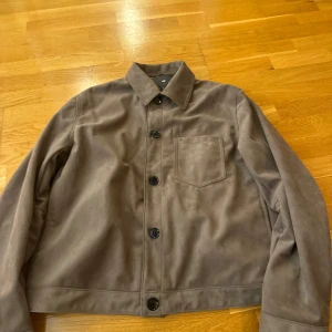 Brun mockajacka från H&M - Snygg brun mockajacka från H&M med knappar framtill och en bröstficka. Perfekt för en stilren look.