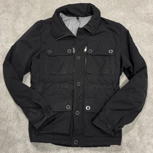 J.lindeberg fieldjacket - Säljer denna svarta Fieldjacket från J.Lindeberg! Jackan är i decent skick, syns att den använts men inga revor, hål eller fläckar, så de finns mycket liv kvar i den! Size L. Skriv vid funderingar!