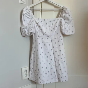Vit klänning med puffärmar från Glamorous - Charmig vit klänning från Glamorous med puffiga kortärmar och ett diskret blommönster. Klänningen har en fyrkantig urringning och en dragkedja i ryggen för enkel påklädning. Perfekt för en somrig look! Klänningen är använd ca 3 gånger därför i nyskick!