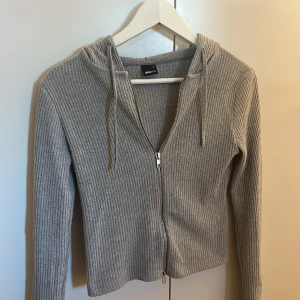 Grå ribbad zip tröja från Gina Tricot - Säljer en stilren grå ribbad zip tröja från Gina Tricot. Den har dragkedja framtill och en justerbar huva med snören. Perfekt för en avslappnad look.