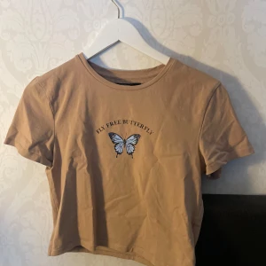 Beige t-shirt med fjärilstryck från Bik Bok - Snygg beige t-shirt från Bik Bok med texten 'Fly Free Butterfly' och ett fjärilstryck på framsidan. Den har en rund halsringning och korta ärmar. Perfekt för en avslappnad stil.