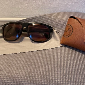 Svarta solglasögon från Ray-Ban - Snygga svarta solglasögon från Ray-Ban med bruna tonade glas. Kommer med ett brunt fodral i läderimitation och en putsduk. Perfekta för soliga dagar och ger en klassisk look.