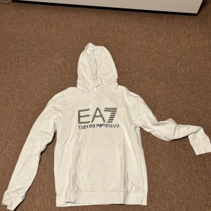 Vit hoodie från Emporio Armani - Säljer en stilren vit hoodie från Emporio Armani med deras ikoniska EA7-logga i svart på bröstet. Tröjan har en klassisk huva med dragsko och en stor magficka framtill. Perfekt för en avslappnad och trendig look.