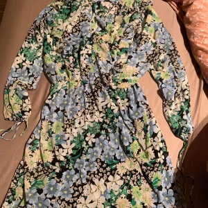 Blommig klänning från H&M - Säljer en färgglad blommig klänning från H&M med långa ärmar och knytband i midjan. Klänningen har ett vackert mönster i grönt, blått och svart, perfekt för vår och sommar.