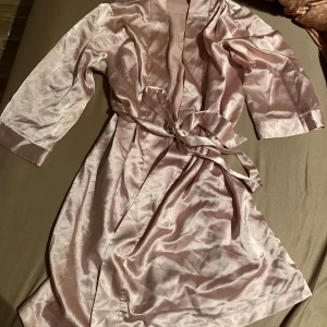Morgonrock  - Elegant ljusrosa morgonrock i siden med glittrande strassdetaljer. Den har trekvartsärmar och ett knytband i midjan för en smickrande passform. Perfekt för en glamorös kväll.