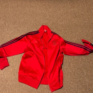 Röd Spanien träningsjacka från Adidas - Snygg röd träningsjacka från Adidas med Spanien logga och blå ränder längs ärmarna e Jackan har dragkedja och ribbade muddar. Perfekt för sportiga aktiviteter.