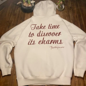 Vit hoodie från Pull&Bear - Säljer en vit hoodie från Pull&Bear med texten 'Take time to discover its charms' i brun på ryggen. Den har en klassisk känguruficka och ribbade muddar. Perfekt för en avslappnad stil.