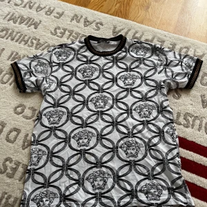 Vit och svart t-shirt från Versace Collection - Snygg vit t-shirt från Versace Collection med svart mönster av Medusa-huvuden och geometriska cirklar. T-shirten har korta ärmar och svarta detaljer vid krage och ärmslut.