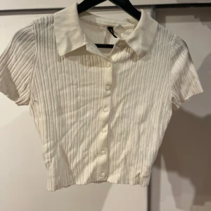 Vit ”ribbad” kortärmad topp - Säljer en stilren vit ribbad topp med korta ärmar och knappar framtill. Perfekt för en avslappnad men ändå elegant look. Toppen har en klassisk krage och är croppad för en modern touch.