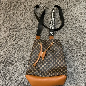 Brun och svart axelväska med mönster - Snygg axelväska i brunt och svart med ett rutigt mönster. Väskan har en justerbar axelrem med text och en praktisk dragsko för stängning. Perfekt för en trendig look.