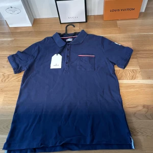  pikétröja från Moncler - Säljer en helt ny stilren mörkblå pikétröja från Moncler i strl large. Tröjan har en liten bröstficka med röd och vit detalj samt en diskret logga på ärmen. Perfekt för en avslappnad men ändå elegant look.