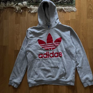 Grå hoodie från Adidas - Säljer en grå hoodie från Adidas med den klassiska röda loggan på framsidan. Tröjan har en stor ficka framtill och en bekväm huva. Perfekt för en avslappnad stil.