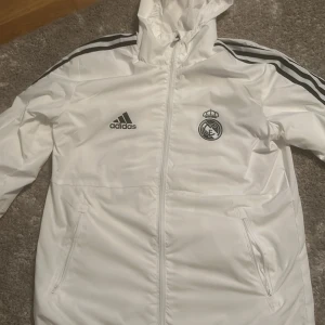 Vit vindjacka från Adidas med Real Madrid-logga - Snygg vit vindjacka från Adidas med Real Madrid-logga på bröstet. Jackan har en huva och dragskedja. Har aldrig använt den så den är i toppen skick. Jackan är i storlek S