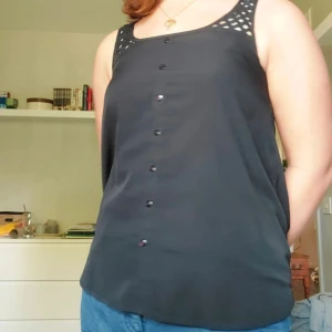 Linne - Ett svart linne med blåa undertoner rutiga axelband. Knappar som ditaljer på frasidan men de går ej att öppna. Tvättlappen är kortklippt men annats i bra skick. Angiven storlek Xs