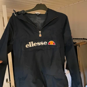Mörkblå vindjacka från Ellesse - Säljer en snygg mörkblå vindjacka från Ellesse. Den har en halv dragkedja framtill och ett stort Ellesse-märke på bröstet. Jackan har långa ärmar och en cool patch på ena ärmen. Perfekt för höst och vår!