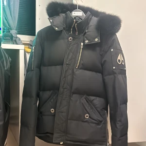 Svart dunjacka från Moose Knuckles - Säljer en svart dunjacka från Moose Knuckles i storlek M. Jackan har en pufferstil med en mysig pälsfodrad huva och flera praktiska fickor med dragkedjor. Perfekt för kalla vinterdagar! 🖤