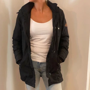 Svart parkajacka med huva - Säljer en svart parkajacka i bra skick. Den har en praktisk huva och stängs med dragkedja och knappar. Perfekt för höst och vår, med en stilren design som passar till det mesta. Jackan har långa ärmar och flera fickor för extra förvaring.