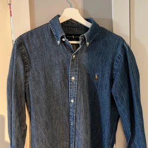 Blå jeansskjorta från Ralph Lauren - Säljer en snygg blå jeansskjorta från Ralph Lauren i storlek S. Skjortan har en klassisk krage och långa ärmar, perfekt för både vardag och fest. Den är i slim fit och har en broderad logga på bröstet. Knapparna framtill ger en stilren look. Passar perfekt till höst och vår!