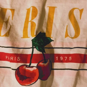 Vit t-shirt med tryck 'Paris 1975' - Säljer en supercool vit t-shirt med tryck 'Paris 1975' och ett par körsbär. Den har en knut framtill för en avslappnad look. Perfekt för sommardagar eller en chill dag på stan. 🏙️