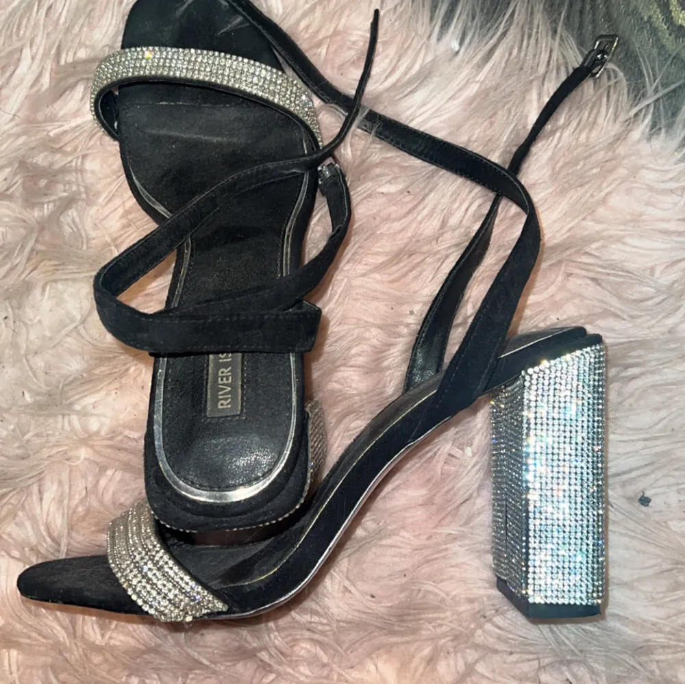 Säljer ett par svarta klackskor från River Island med glittriga remmar och en rejäl klack täckt av strass. Perfekta för en festkväll när du vill sticka ut! Skorna har en elegant design med en smal rem runt ankeln för extra stöd.. Kengät.