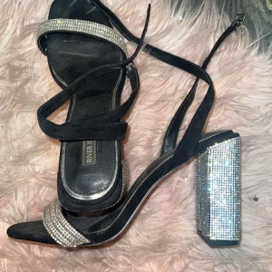 Svarta klackskor med glittriga detaljer från River Island - Säljer ett par svarta klackskor från River Island med glittriga remmar och en rejäl klack täckt av strass. Perfekta för en festkväll när du vill sticka ut! Skorna har en elegant design med en smal rem runt ankeln för extra stöd.