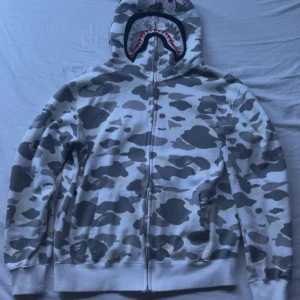 Grå camo hoodie från Bape - Säljer en Bape Hoodie med haj, i mycket bra skick  och håller dig varm under höst och vår. Storlek XL men Passar mer L än XL,säljer den pga har ingen användning av den mer
