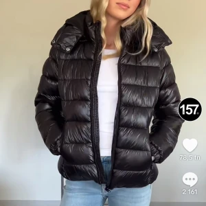 Svart pufferjacka - Säljer denna svarta trendiga pufferjacka som är perfekt för kyliga dagar. Jackan har en glansig finish och dragkedja framtill. Den är långärmad och har en mysig huva. Perfekt för både höst och vinter ❄️