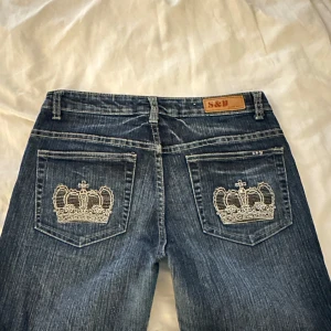 Blå jeans med broderade kronor - Snygga blå jeans från S&Y med unika broderade kronor på bakfickorna. De har en cool bootcut-stil och är dekorerade med glittriga detaljer framtill. Perfekta för att ge din outfit en extra touch av stil. Passar både till vardags och fest! Midja: 36cm och innerben: 70cm 