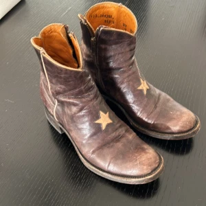 Bruna boots med stjärndetalj - Säljer ett par coola mexikana  bruna boots i skinn med en snygg stjärndetalj på framsidan. De har en dragkedja på sidan för enkel på- och avtagning. Perfekta för höst och vår, dessa boots ger en unik touch till din outfit. Använda men i bra skick.