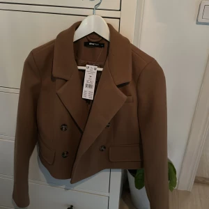 Beige kavaj/jacka från Gina Tricot - Säljer en snygg beige kavaj från Gina Tricot i croppad modell. Den har en dubbelknäppt design med stiliga knappar och långa ärmar. Perfekt för höst och vår, och funkar både till vardags och fest. Kavajen är i nytt skick med prislappen kvar. 🧥✨